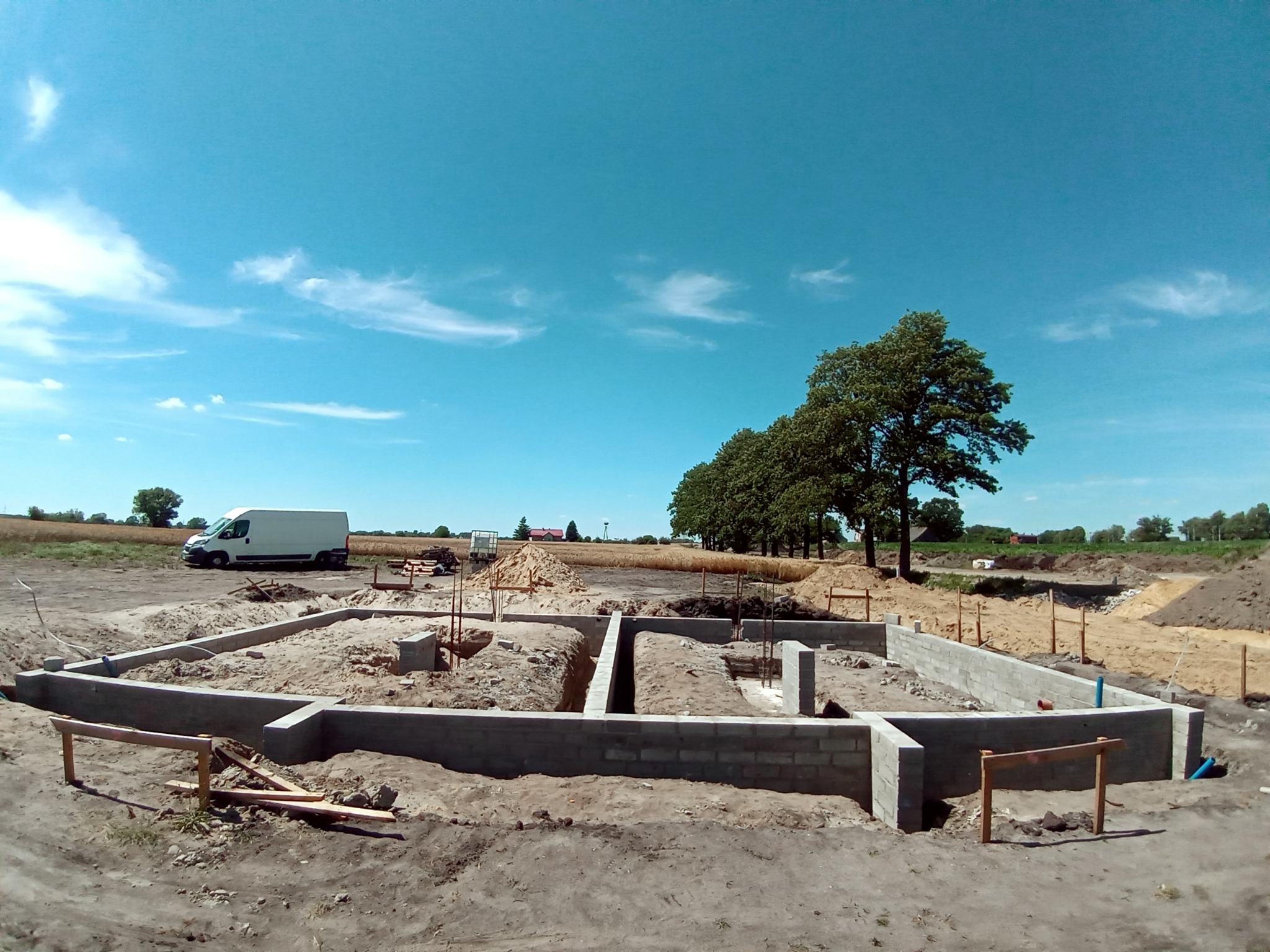 Wylane fundamenty pod budynek z widocznymi zbrojeniami, ujęcie w słoneczny dzień z perspektywy szerokokątnej, na horyzoncie widoczny krajobraz polny i biały samochód dostawczy.