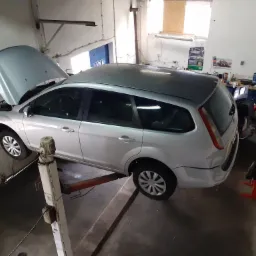 Srebrny samochód kombi na podnośniku w warsztacie, z otwartą maską i widocznym silnikiem, otoczenie warsztatowe z narzędziami i plakatami na ścianach.