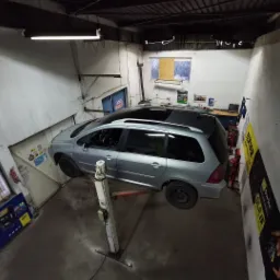 Srebrny samochód typu kombi na podnośniku w warsztacie, widoczne plakaty reklamowe na ścianie i narzędzia w tle.