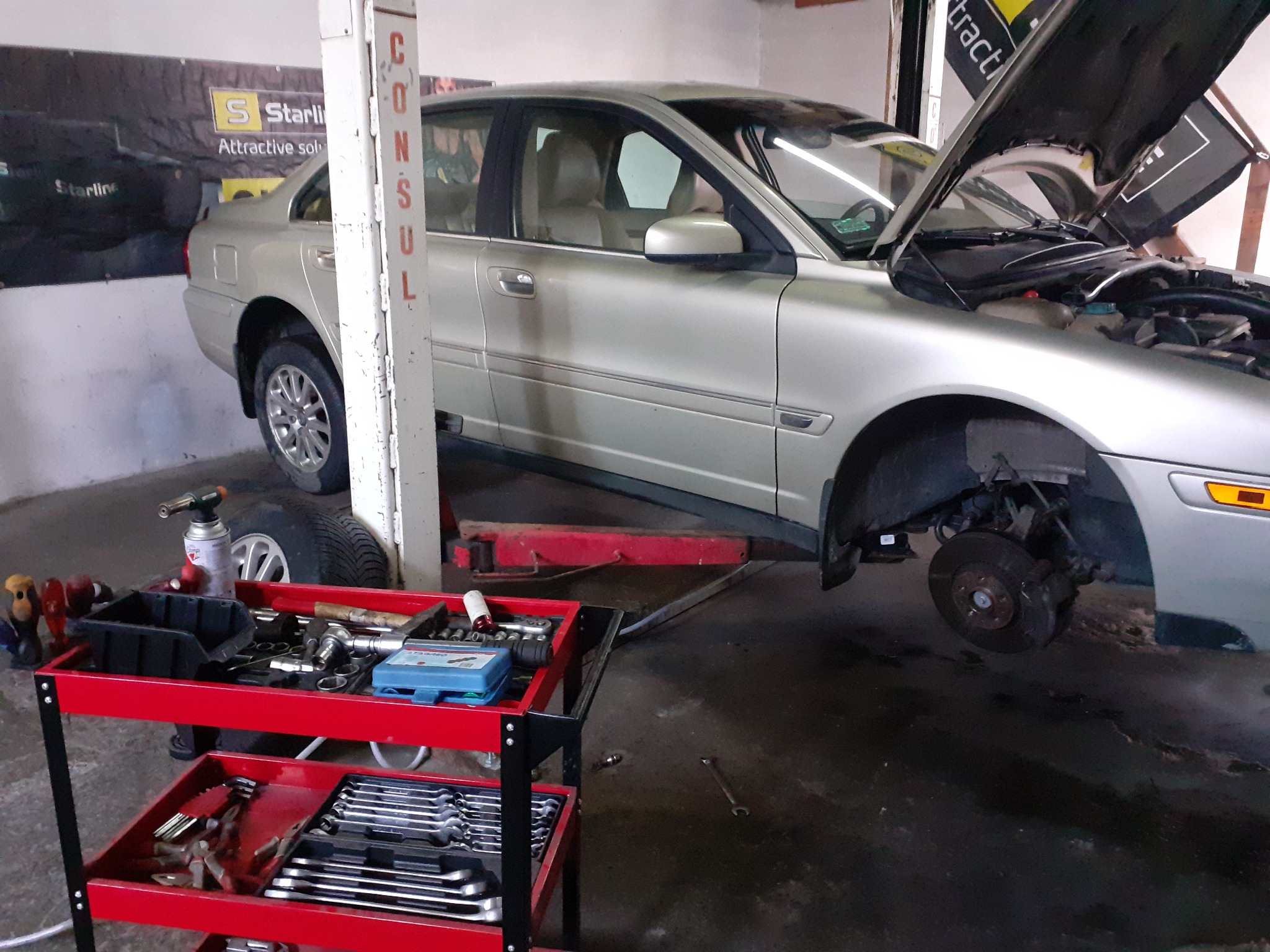 Srebrny samochód z otwartą maską, podniesiony na podnośniku, z widocznym brakiem koła, na tle warsztatu z wózkiem narzędziowym i reklamą na ścianie.