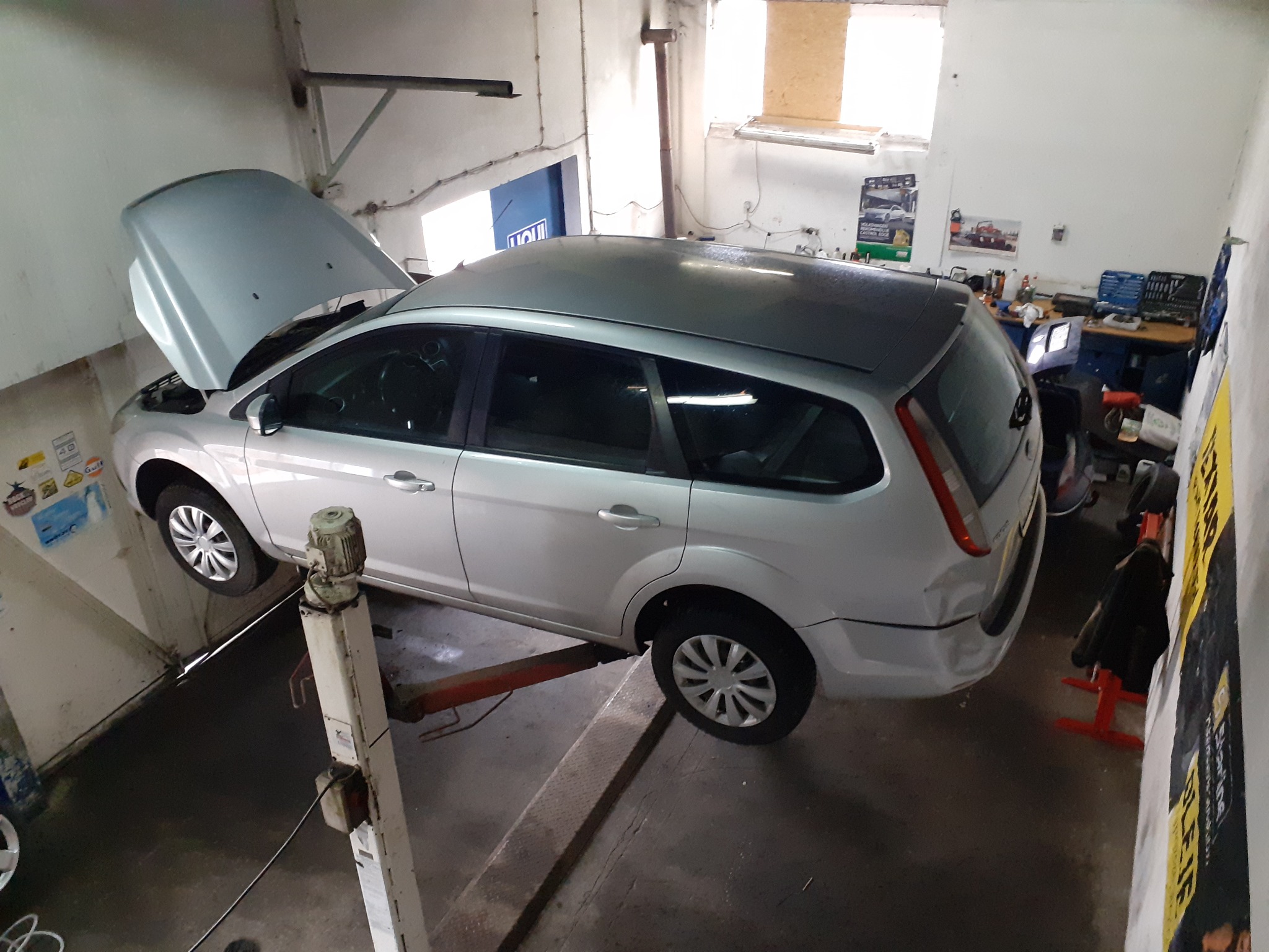 Srebrny samochód kombi na podnośniku w warsztacie, z otwartą maską i widocznym silnikiem, otoczenie warsztatowe z narzędziami i plakatami na ścianach.