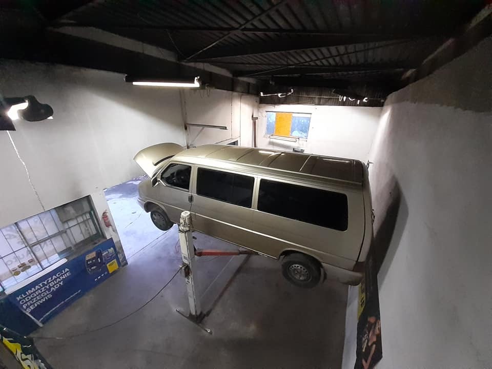 Złoty van Volkswagen T4 na podnośniku w warsztacie samochodowym z otwartą maską, widok z góry, widoczne plakaty reklamowe na ścianie.