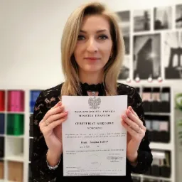 Kobieta z blond włosami prezentuje certyfikat księgowego wydany przez Ministra Finansów Rzeczypospolitej Polskiej, na tle półki z segregatorami i czarno-białymi fotografiami.