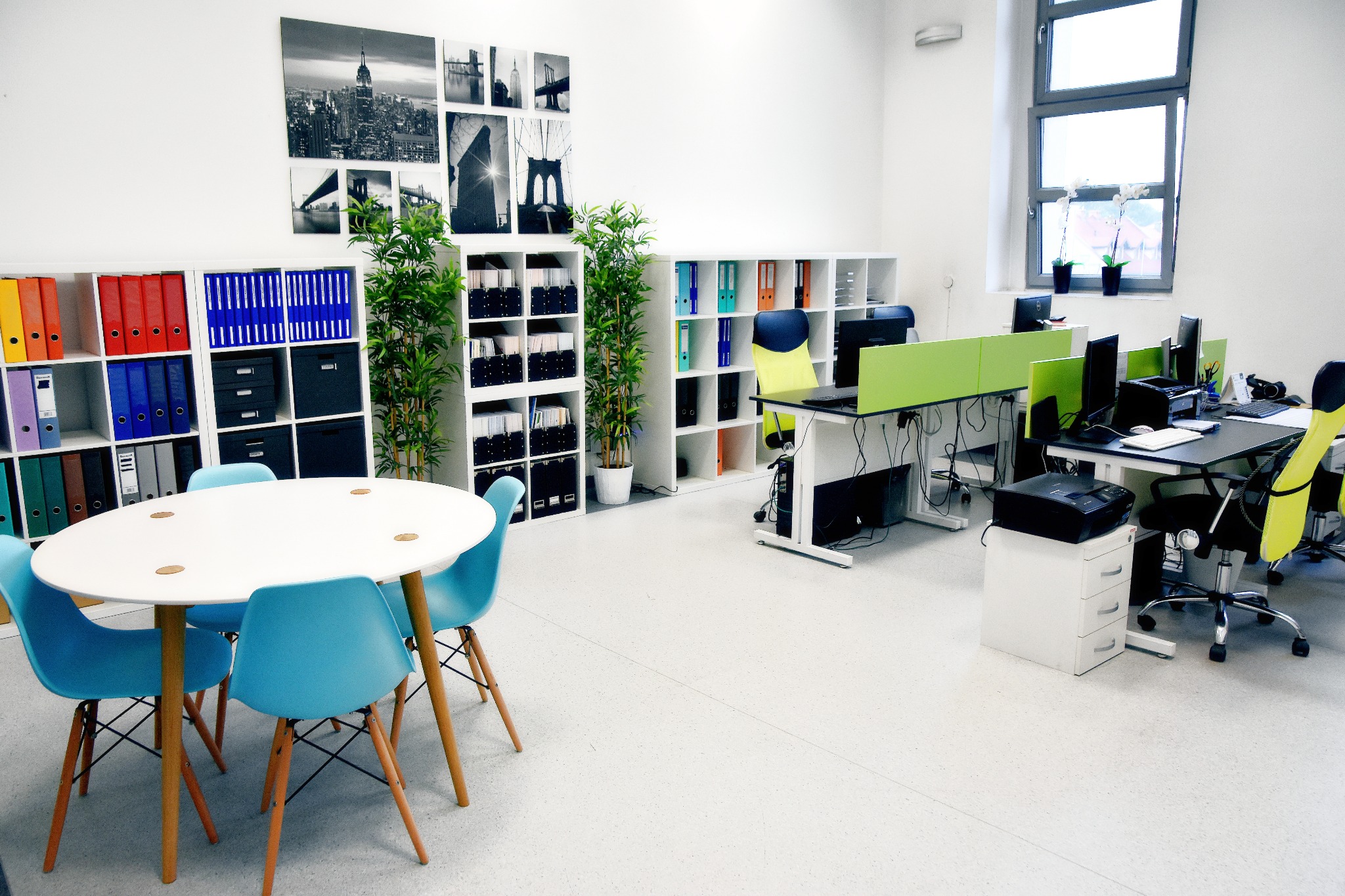 Jasne, przestronne biuro z białymi regałami wypełnionymi kolorowymi segregatorami, biurkami z zielonymi przegrodami i okrągłym stołem z turkusowymi krzesłami.