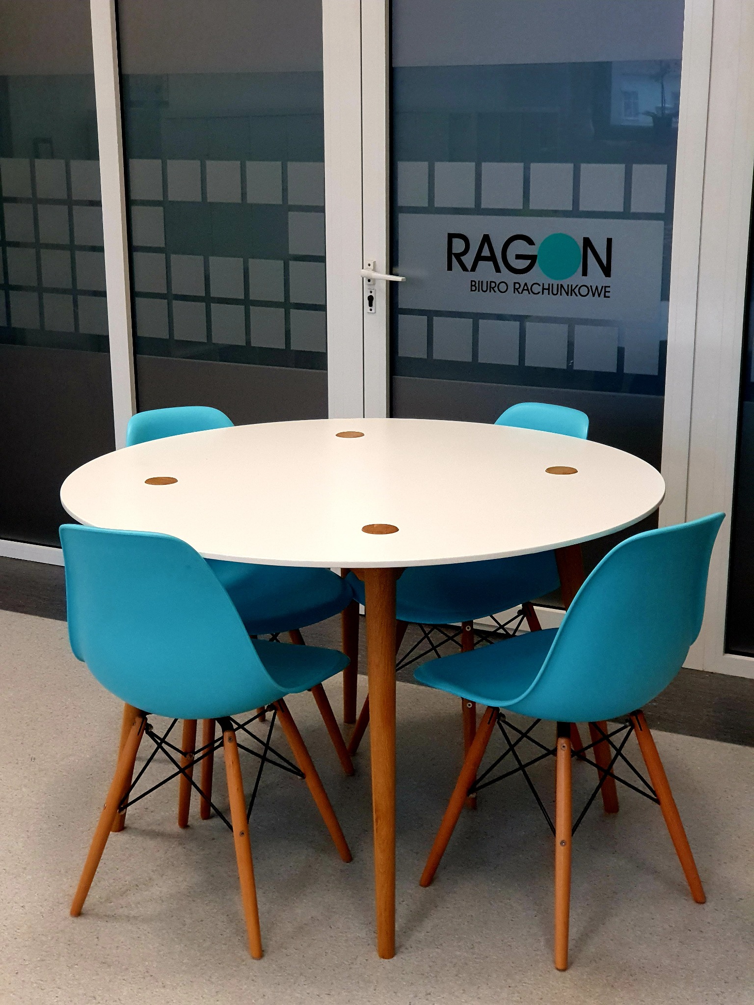 Okrągły biały stół z czterema turkusowymi krzesłami w minimalistycznym biurze, logo firmy na szklanej ścianie w tle.
