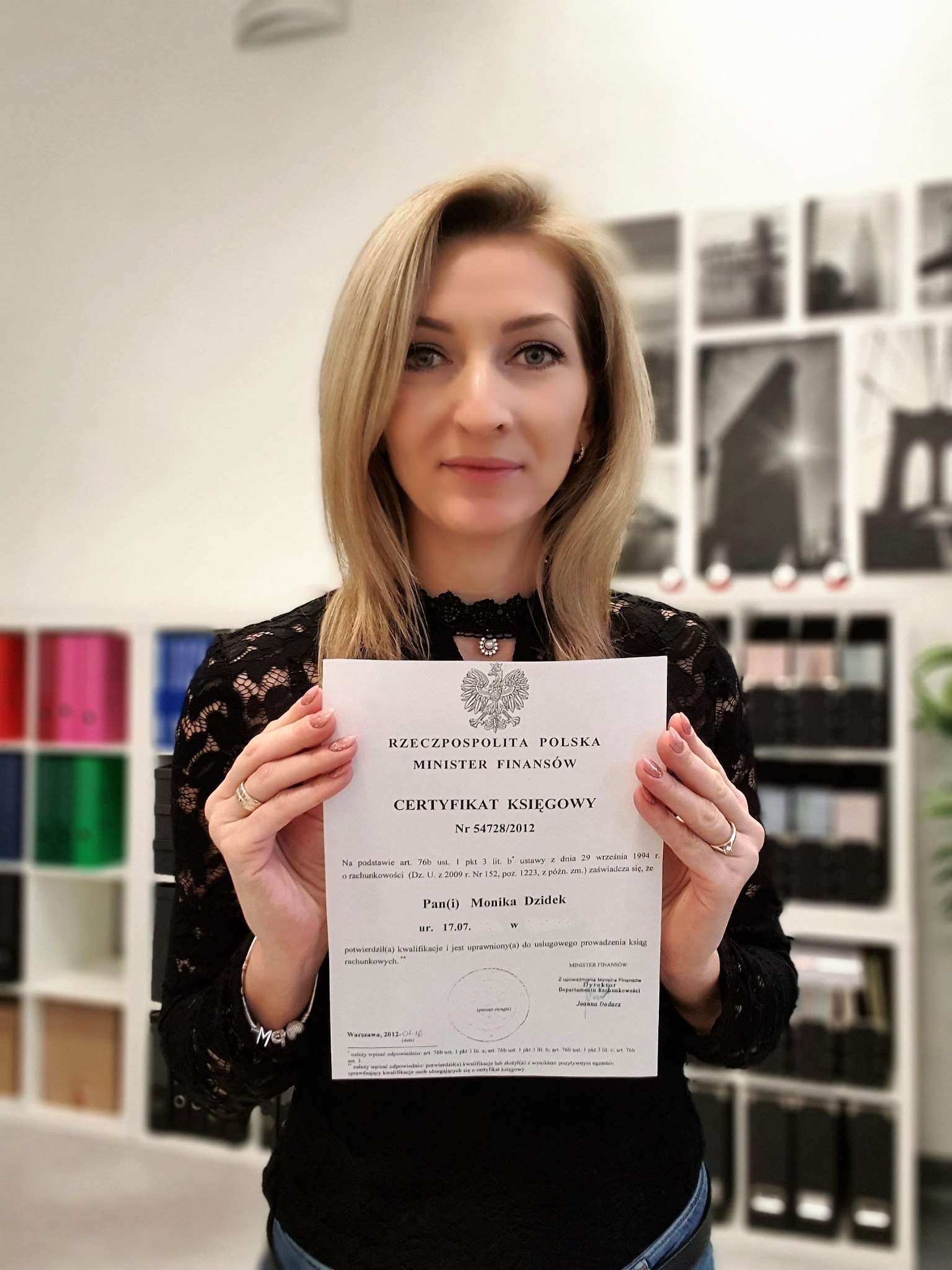 Kobieta z blond włosami prezentuje certyfikat księgowego wydany przez Ministra Finansów Rzeczypospolitej Polskiej, na tle półki z segregatorami i czarno-białymi fotografiami.