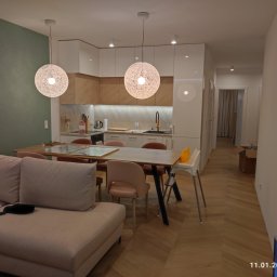 Dom Basen Ogród Marcin Kłos - Nowoczesne wnętrze salonu z kuchnią: dwa okrągłe, ażurowe wiszące światła nad drewnianym stołem otoczonym różowymi krzesłami, białe szafki kuchenne z drewnianymi akcentami w tle, oraz beżowa sofa...