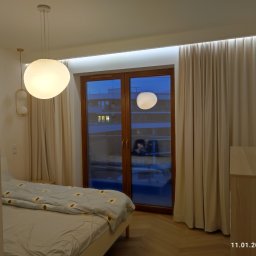 Dom Basen Ogród Marcin Kłos - Sypialnia z dużym oknem, zasłonami, wiszącymi lampami kulistymi i oświetleniem LED nad oknem, widok na budynek z zewnątrz.