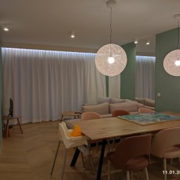 Dom Basen Ogród Marcin Kłos - Jasne wnętrze salonu z drewnianym stołem i krzesłami, oświetlone dwoma wiszącymi lampami z włókna, parkiet w jodełkę na podłodze i minimalistyczny wystrój.