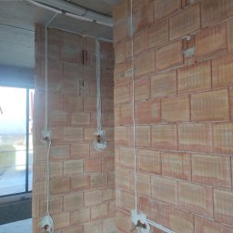 Dom Basen Ogród Marcin Kłos - Wewnętrzne ściany z czerwonej cegły z bruzdami pod instalację elektryczną, widoczne kable i puszki, rury ochronne na suficie, stan surowy.