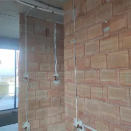 Wewnętrzne ściany z czerwonej cegły z bruzdami pod instalację elektryczną, widoczne kable i puszki, rury ochronne na suficie, stan surowy.