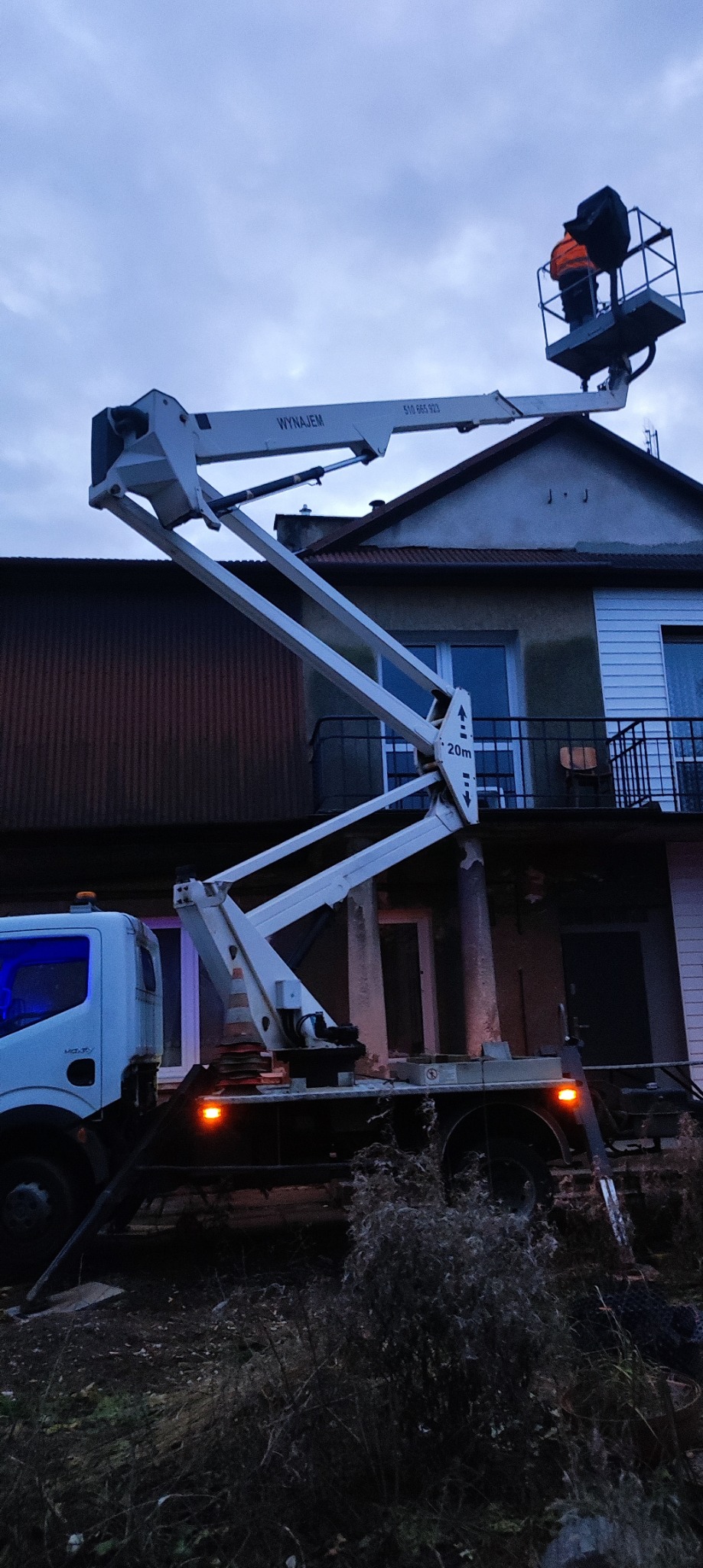 Dwóch pracowników w pomarańczowych kamizelkach na podnośniku koszowym, pracujących przy elewacji dwupiętrowego budynku z balkonem, widoczny fragment ciężarówki z podnośnikiem na tle pochmurnego nieba.