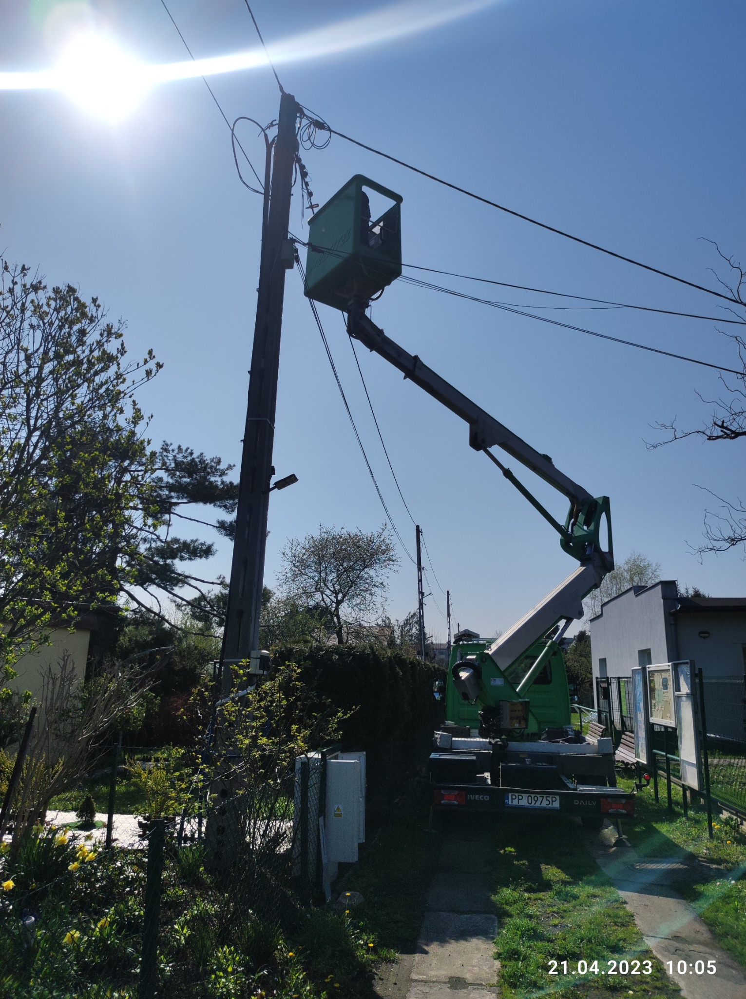 Pracownik w podnośniku koszowym przy słupie energetycznym, widoczne linie wysokiego napięcia, drzewa, budynek i fragment ogrodzenia, słoneczny dzień.