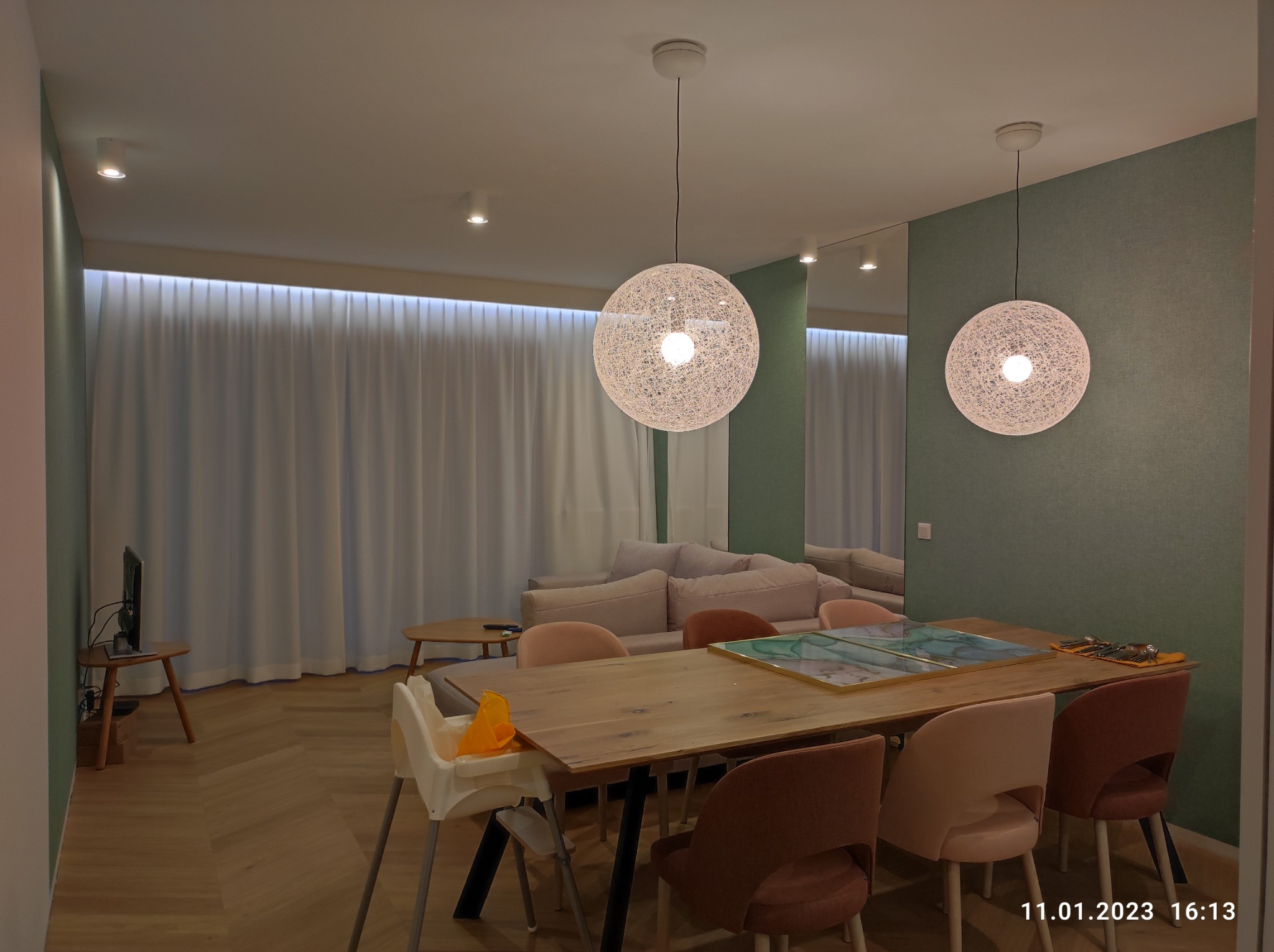 Jasne wnętrze jadalni z drewnianym stołem, krzesłami w odcieniach różu i brązu, oraz dwoma wiszącymi lampami kulistymi z plecionki; w tle zasłonięte okno i kanapa.