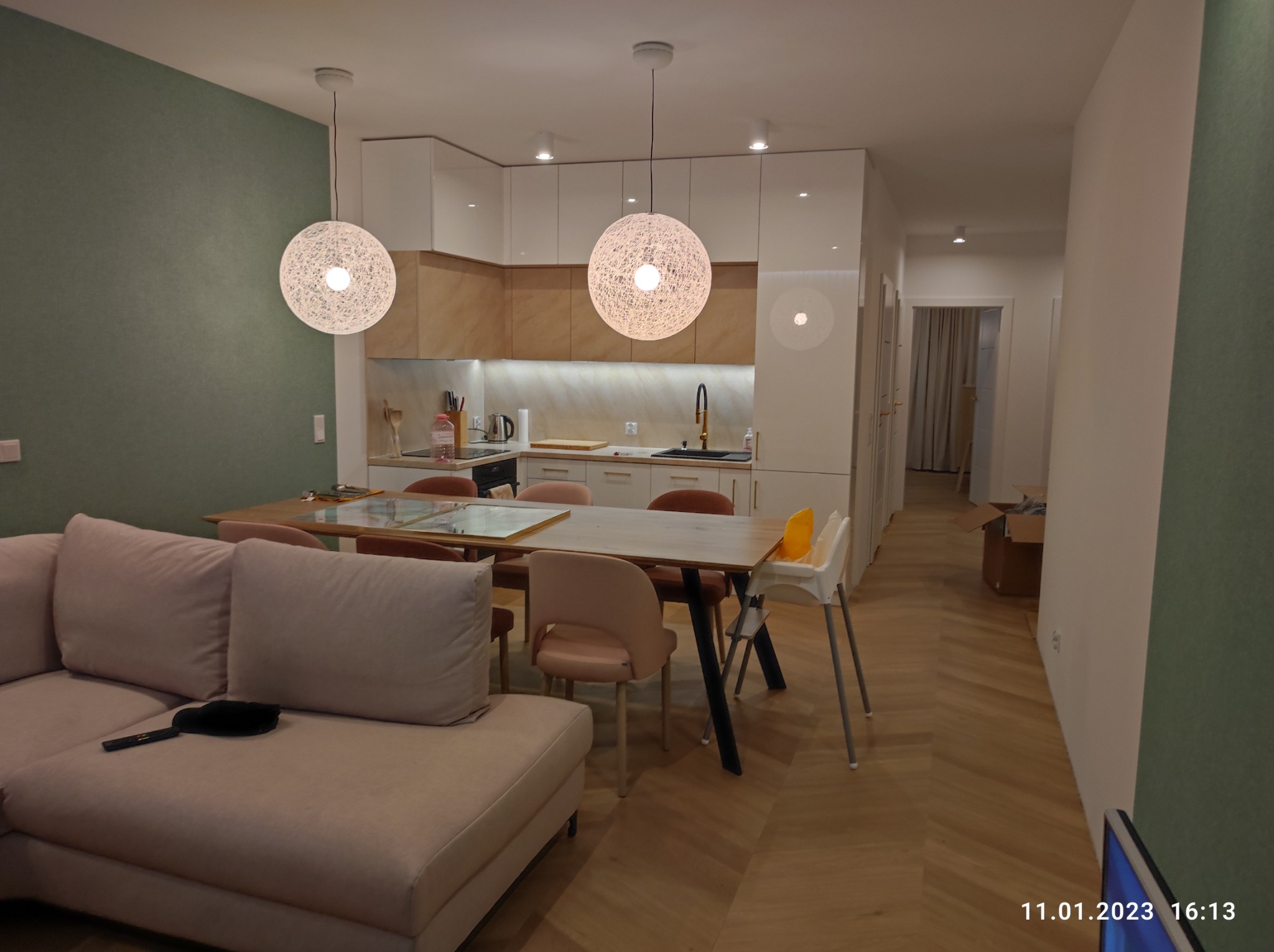 Nowoczesne wnętrze salonu z kuchnią: dwa okrągłe, ażurowe wiszące światła nad drewnianym stołem otoczonym różowymi krzesłami, białe szafki kuchenne z drewnianymi akcentami w tle, oraz beżowa sofa...