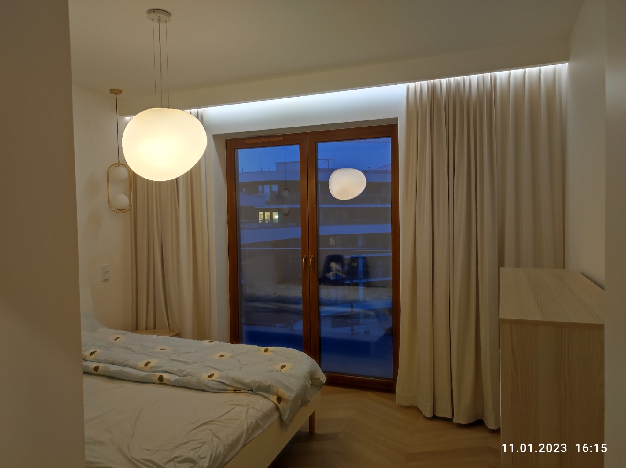Sypialnia z dużym oknem, zasłonami, wiszącymi lampami kulistymi i oświetleniem LED nad oknem, widok na budynek z zewnątrz.