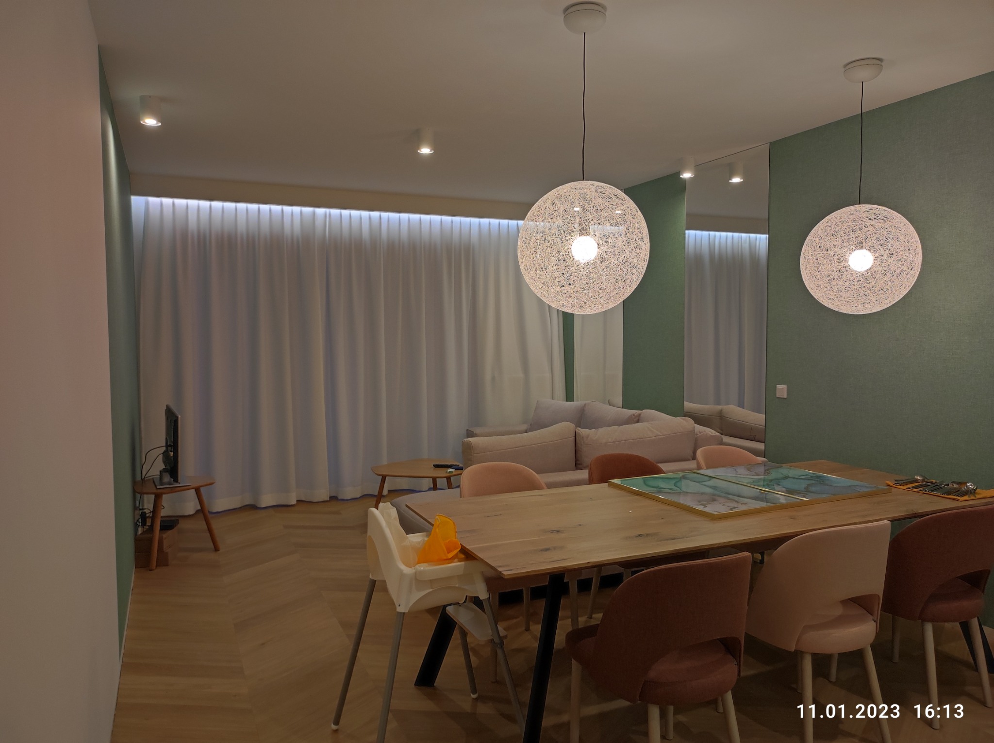 Jasne wnętrze salonu z drewnianym stołem i krzesłami, oświetlone dwoma wiszącymi lampami z włókna, parkiet w jodełkę na podłodze i minimalistyczny wystrój.