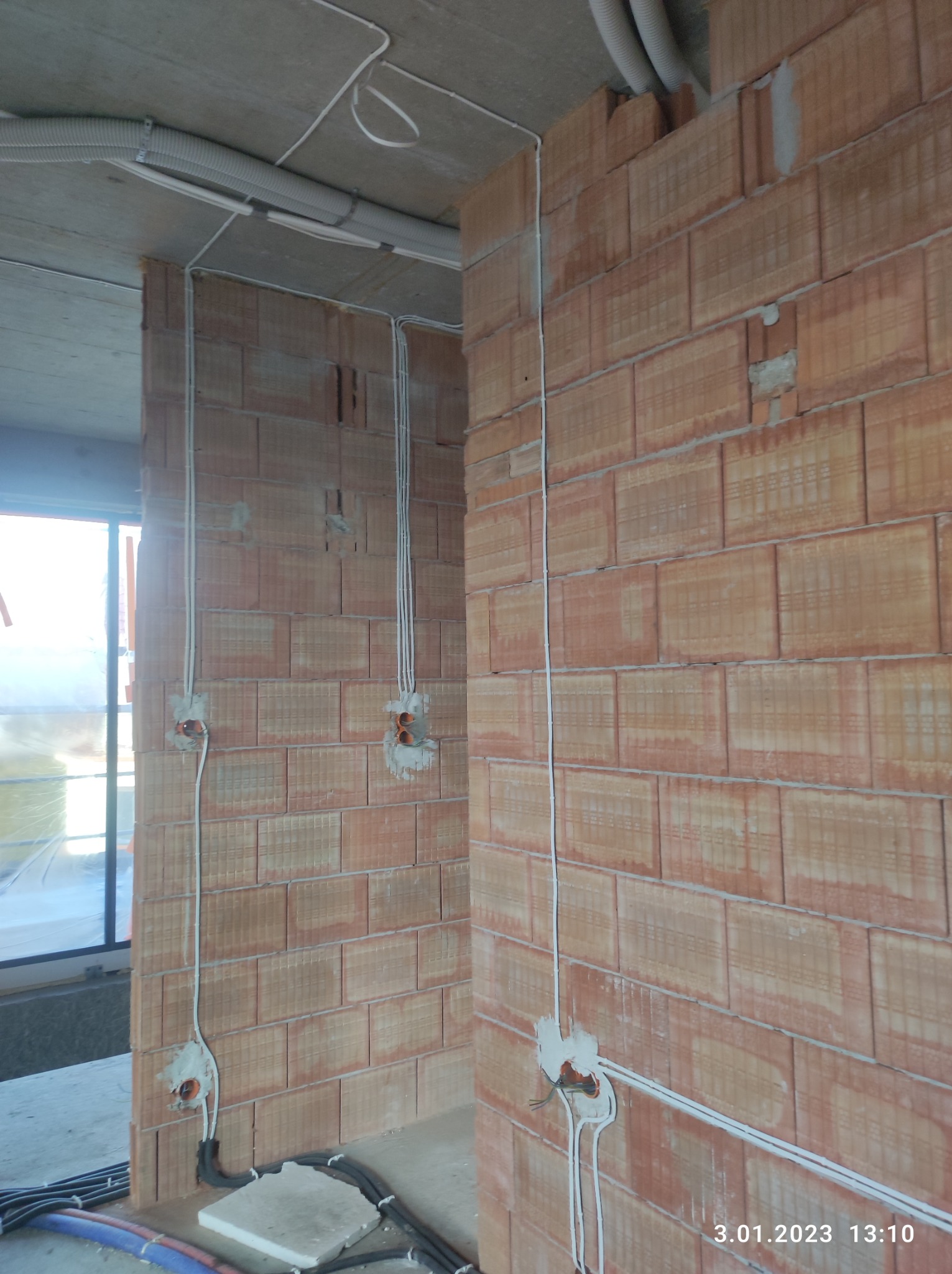 Wewnętrzne ściany z czerwonej cegły z bruzdami pod instalację elektryczną, widoczne kable i puszki, rury ochronne na suficie, stan surowy.