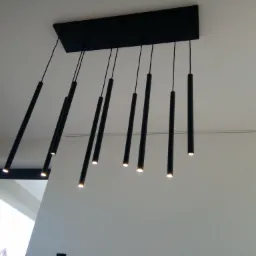 Czarna, podłużna lampa sufitowa z ośmioma cylindrycznymi, wiszącymi elementami świecącymi ciepłym światłem, widoczna na tle białego sufitu i fragmentu okna.