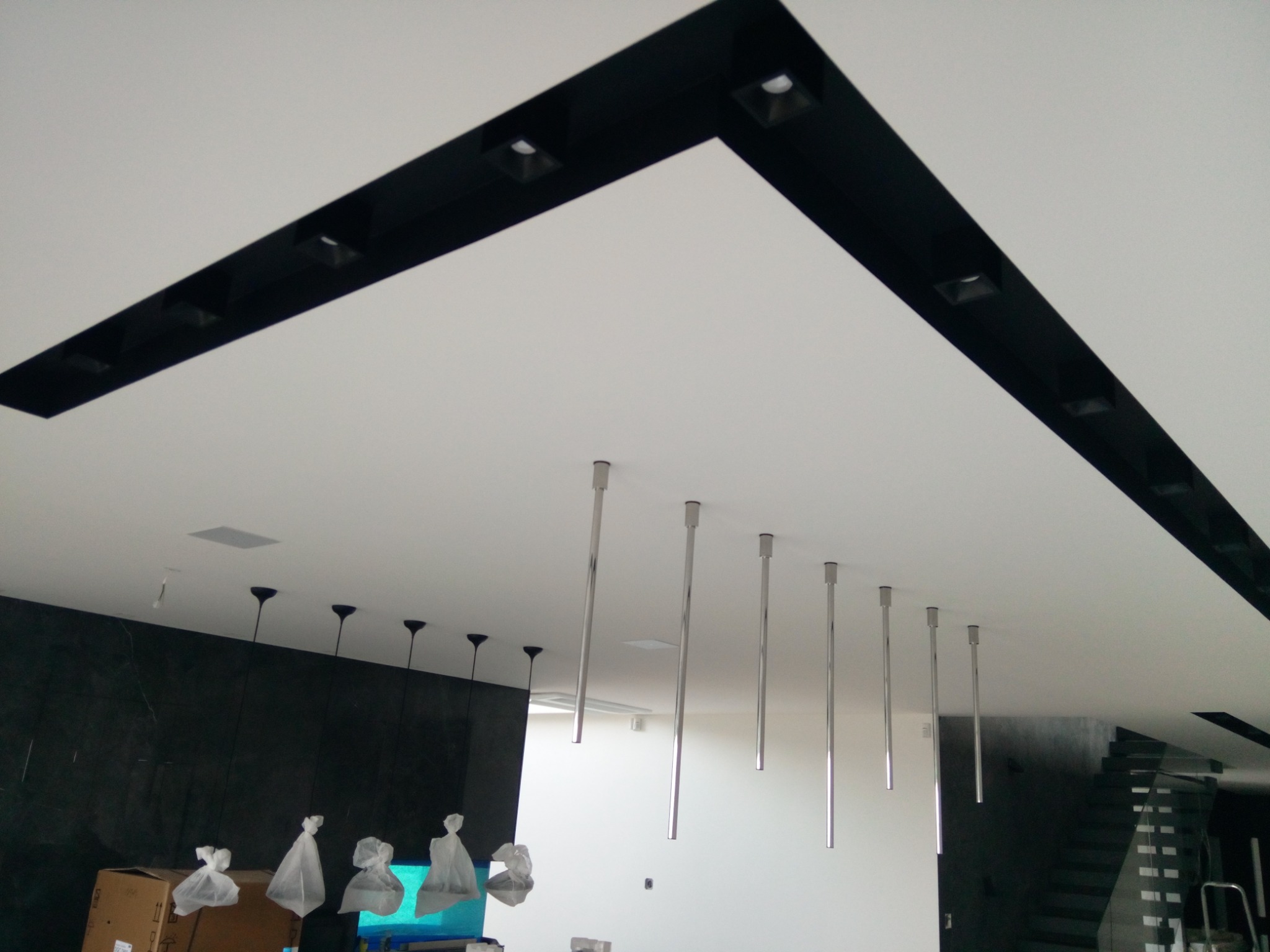Nowoczesny sufit z czarnymi, geometrycznymi listwami i wbudowanymi reflektorami, uzupełniony o metalowe, cylindryczne lampy wiszące na długich prętach, niektóre jeszcze w folii ochronnej.