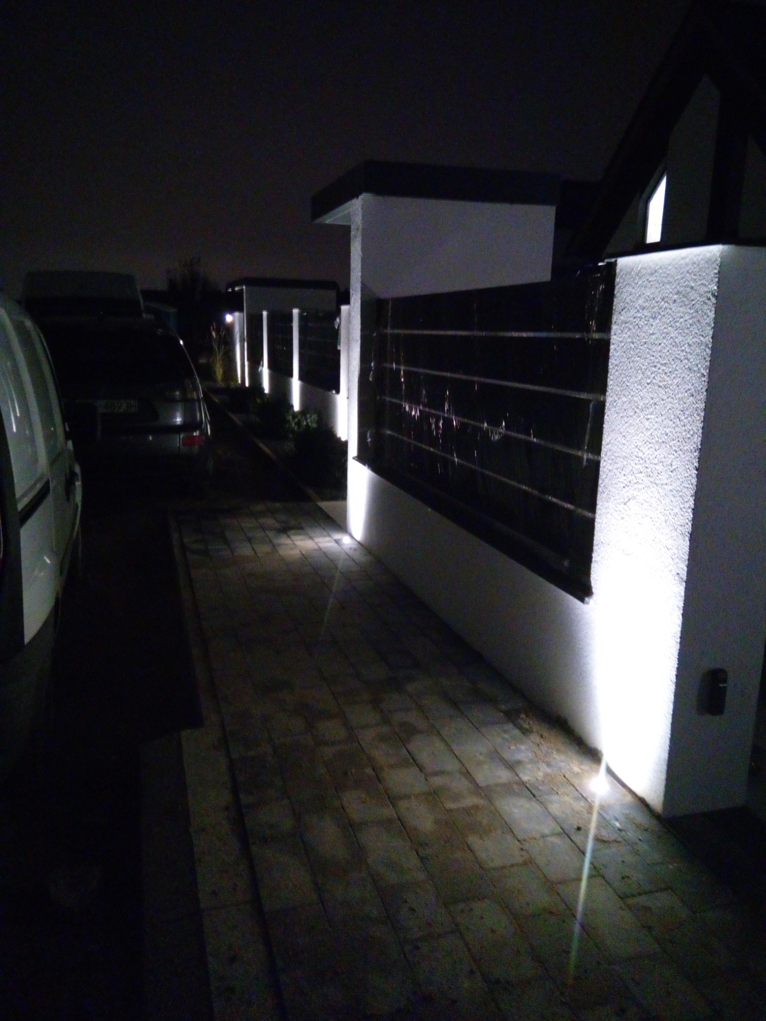 Oświetlenie ścieżki i ogrodzenia posesji nocą, z widocznymi lampami wbudowanymi w kostkę brukową i słupki ogrodzeniowe.