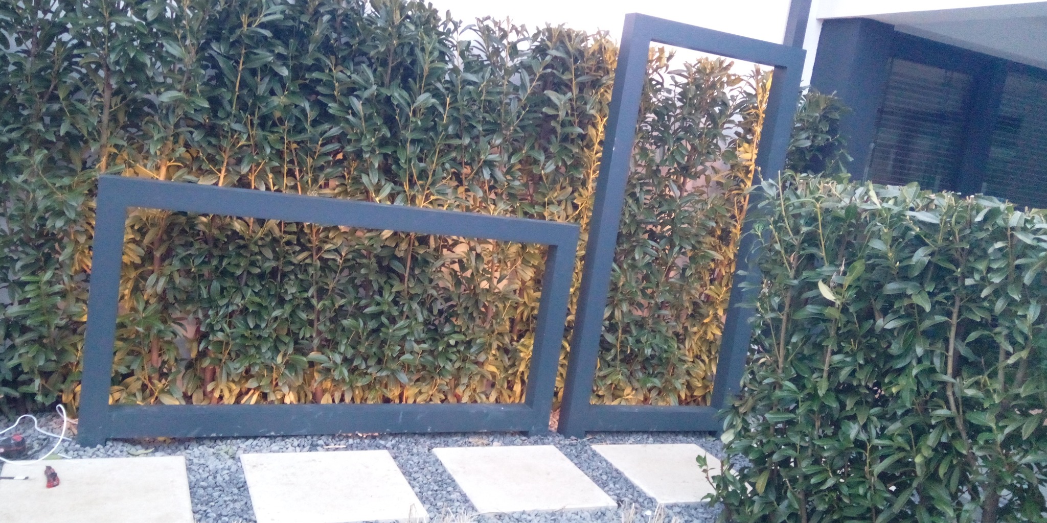 Nowoczesna aranżacja ogrodu z podświetlanym żywopłotem i geometrycznymi ramami z przewodami elektrycznymi widocznymi w tle.