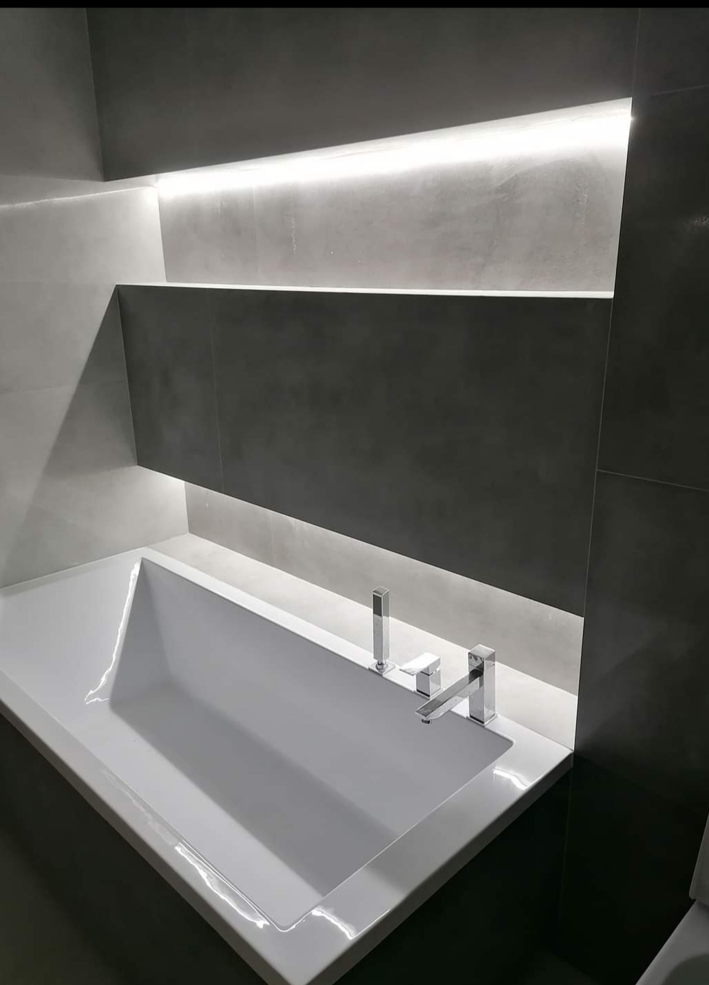 Nowoczesna łazienka z białą wanną w zabudowie, szare płytki i podświetlana półka wnękowa, minimalistyczna armatura.