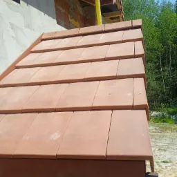 Nowo położony dach z terakoty z widoczną rynną, fragmentem elewacji w trakcie budowy i zielenią w tle.