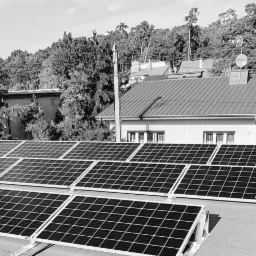 Czarno-białe zdjęcie instalacji paneli słonecznych na dachu budynku mieszkalnego, widok z góry, tło zalesione.