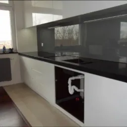 Nowoczesna kuchnia z białymi szafkami, czarnym blatem i szarym panelem szklanym między szafkami, widoczne rury odpływowe pod zlewem.