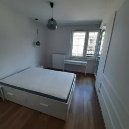 Jasne wnętrze sypialni z drewnianą podłogą, białym łóżkiem z szufladami, biurkiem pod oknem i minimalistycznymi lampami wiszącymi.