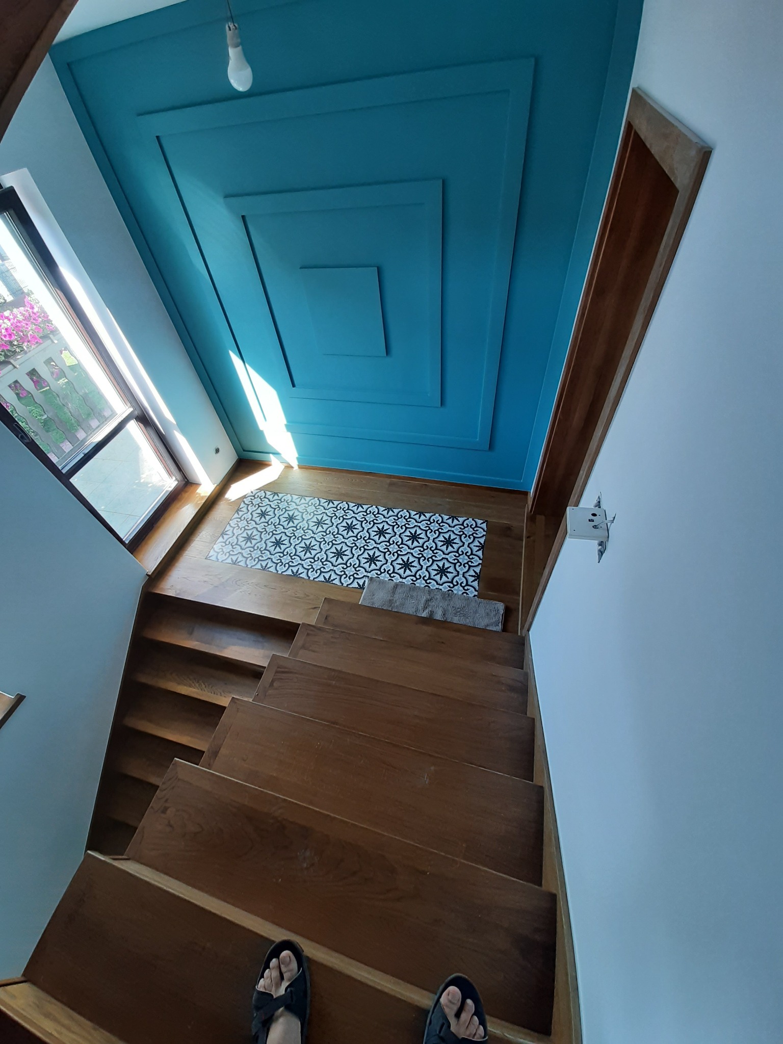 Widok z góry na drewniane schody z ciemnymi stopniami, prowadzące w dół do przedpokoju z wzorzystą, czarno-białą podłogą, turkusowy sufit z kasetonami i widokiem na okno z kwiatami.