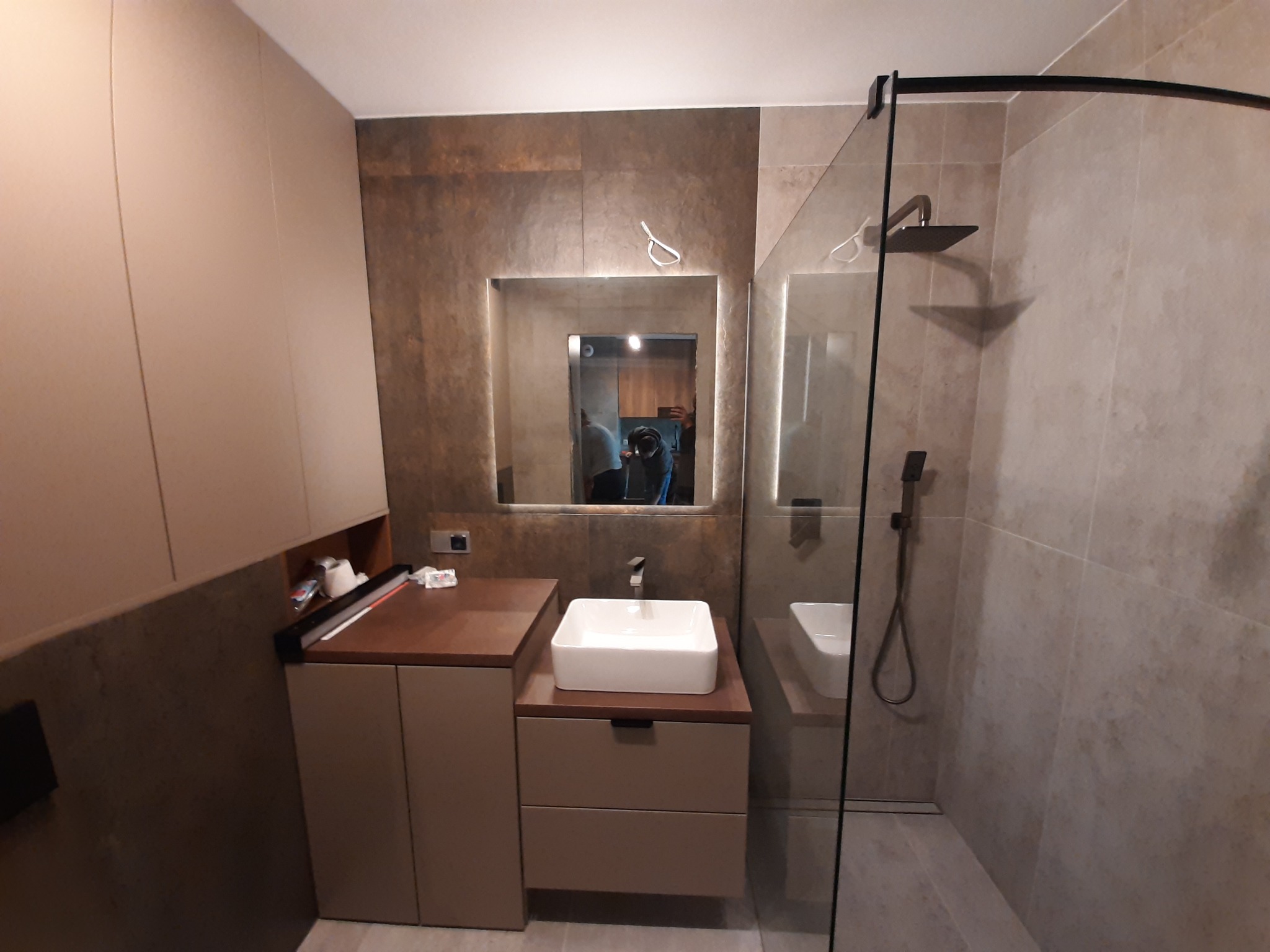 Nowoczesna łazienka w stonowanych brązach i beżach, z prostokątną umywalką nablatową, podświetlanym lustrem i kabiną prysznicową typu walk-in z czarnymi akcentami.