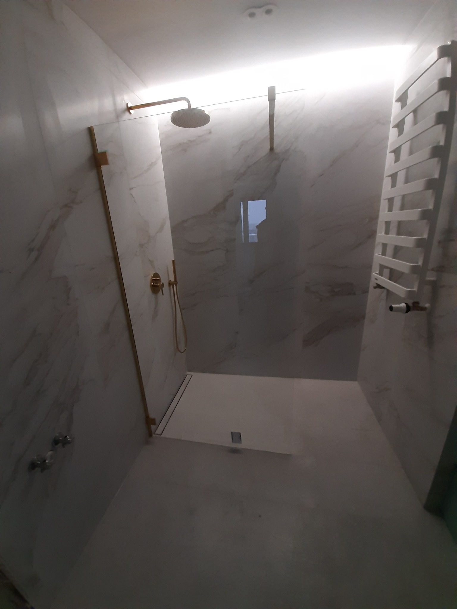 Nowoczesna kabina prysznicowa walk-in z marmurowymi płytkami, złotą armaturą i białym grzejnikiem drabinkowym.