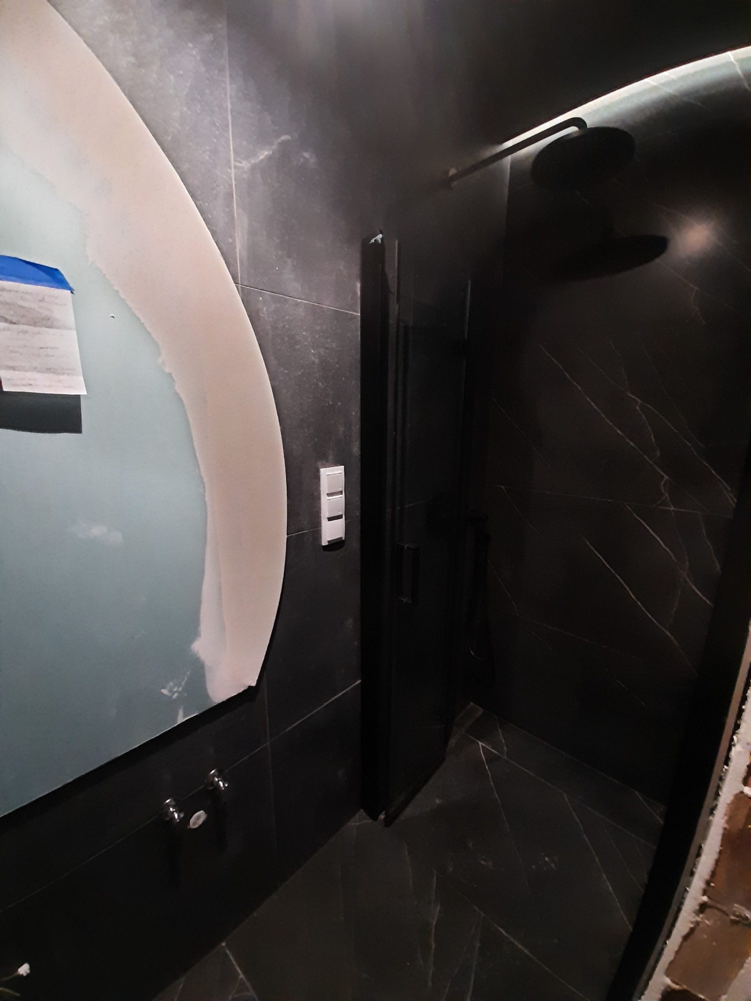 Ciemna łazienka w trakcie wykańczania, widoczna owalna rama lustra z nałożoną szpachlą, czarne płytki imitujące marmur, kabina prysznicowa z czarnymi elementami i biały włącznik światła.