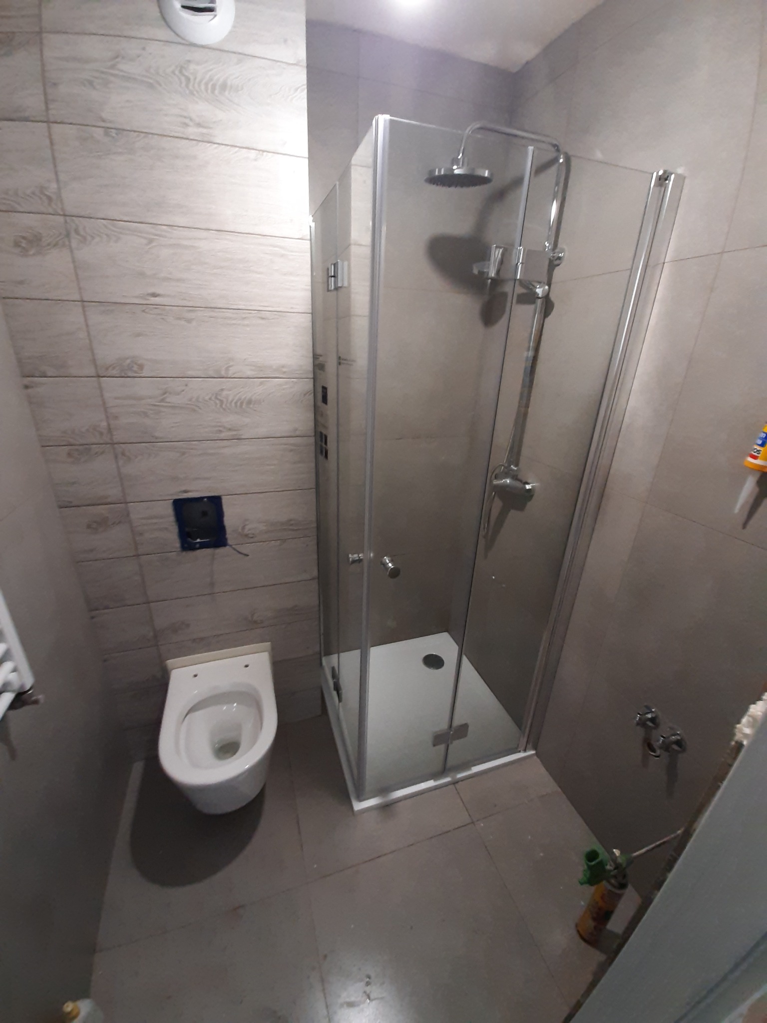 Nowoczesna łazienka po remoncie: kabina prysznicowa z chromowanym zestawem natryskowym, podwieszana toaleta z zabudowanym stelażem, płytki imitujące drewno na ścianach, szara podłoga i puszka...