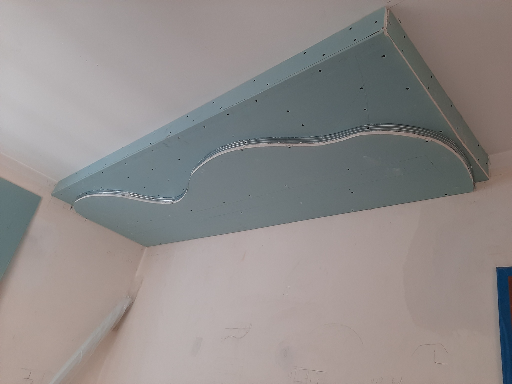 Niezaszpachlowana, podwieszana konstrukcja z płyt gipsowo-kartonowych na suficie, z dekoracyjnym, falistym wycięciem, widoczne ślady ołówka na ścianach.