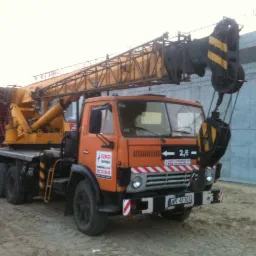 Pomarańczowy dźwig samochodowy marki Jelcz z żółtym wysięgnikiem na placu budowy, widoczne betonowe elementy konstrukcyjne w tle.