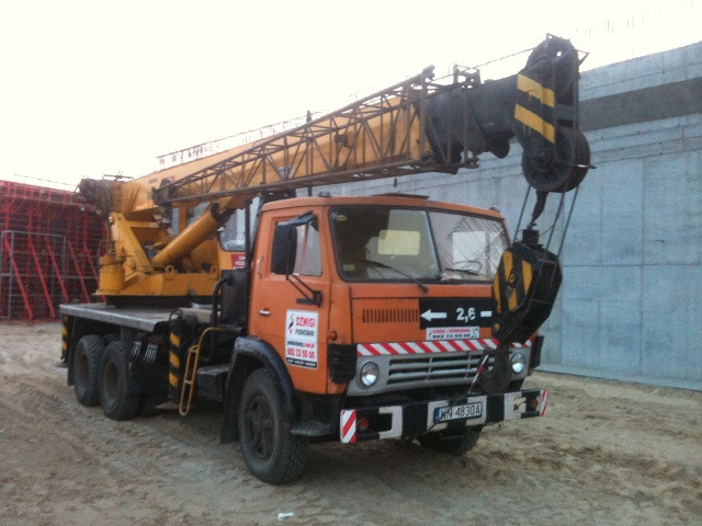 Pomarańczowy dźwig samochodowy marki Jelcz z żółtym wysięgnikiem na placu budowy, widoczne betonowe elementy konstrukcyjne w tle.