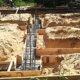 Przygotowanie wykopów i szalunków zbrojeniowych pod fundamenty domu jednorodzinnego na piaszczystej działce, widoczny pracownik w tle.