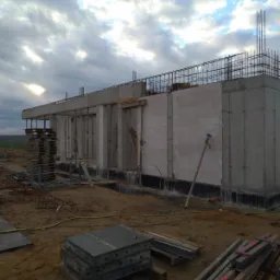 Budowa domu jednorodzinnego w stanie surowym otwartym, widoczne ściany z betonu komórkowego, zbrojenie stropu, podpory szalunkowe i materiały budowlane na placu budowy, pochmurne niebo w tle.