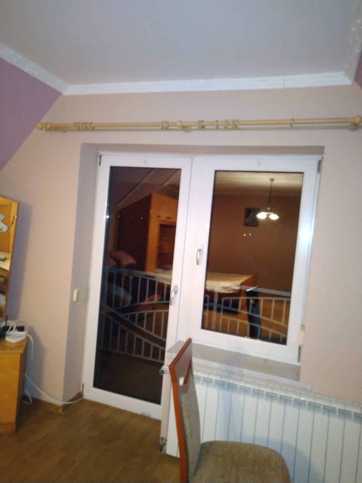 Dwuskrzydłowe okno balkonowe z białymi ramami i widokiem na wnętrze domu z balustradą, grzejnik panelowy pod oknem, drewniany karnisz.