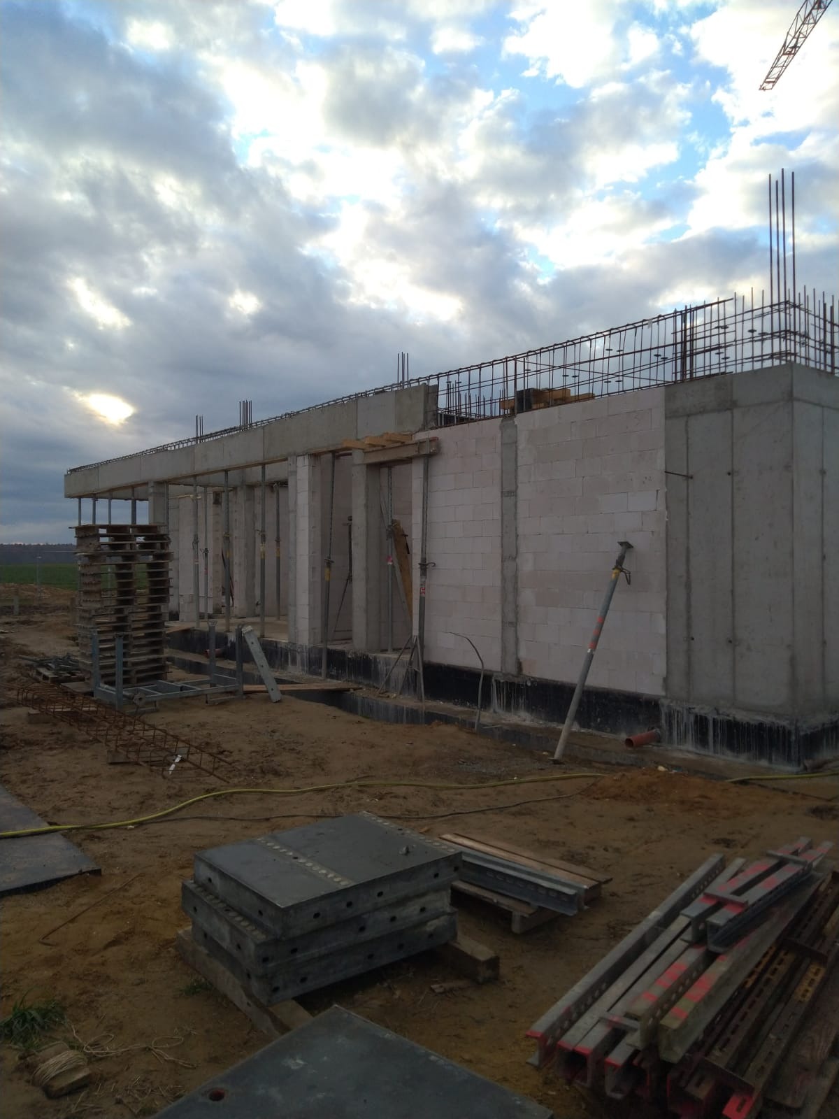 Budowa domu jednorodzinnego w stanie surowym otwartym, widoczne ściany z betonu komórkowego, zbrojenie stropu, podpory szalunkowe i materiały budowlane na placu budowy, pochmurne niebo w tle.