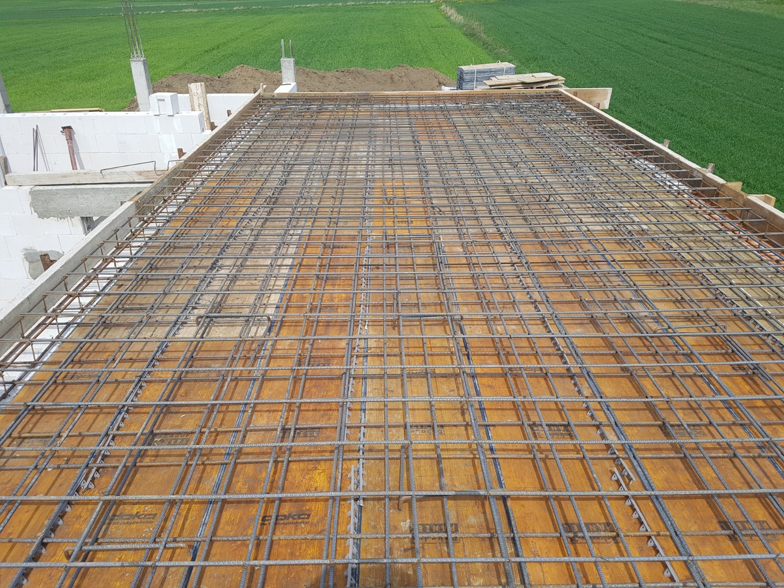Zbrojenie stropu domu jednorodzinnego z widoczną konstrukcją szalunkową i fragmentem muru z betonu komórkowego w tle krajobraz polny.