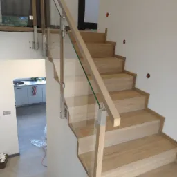 Jasne, drewniane stopnie schodów z szklaną balustradą i metalowymi mocowaniami, widok z dołu, w tle fragment kuchni.