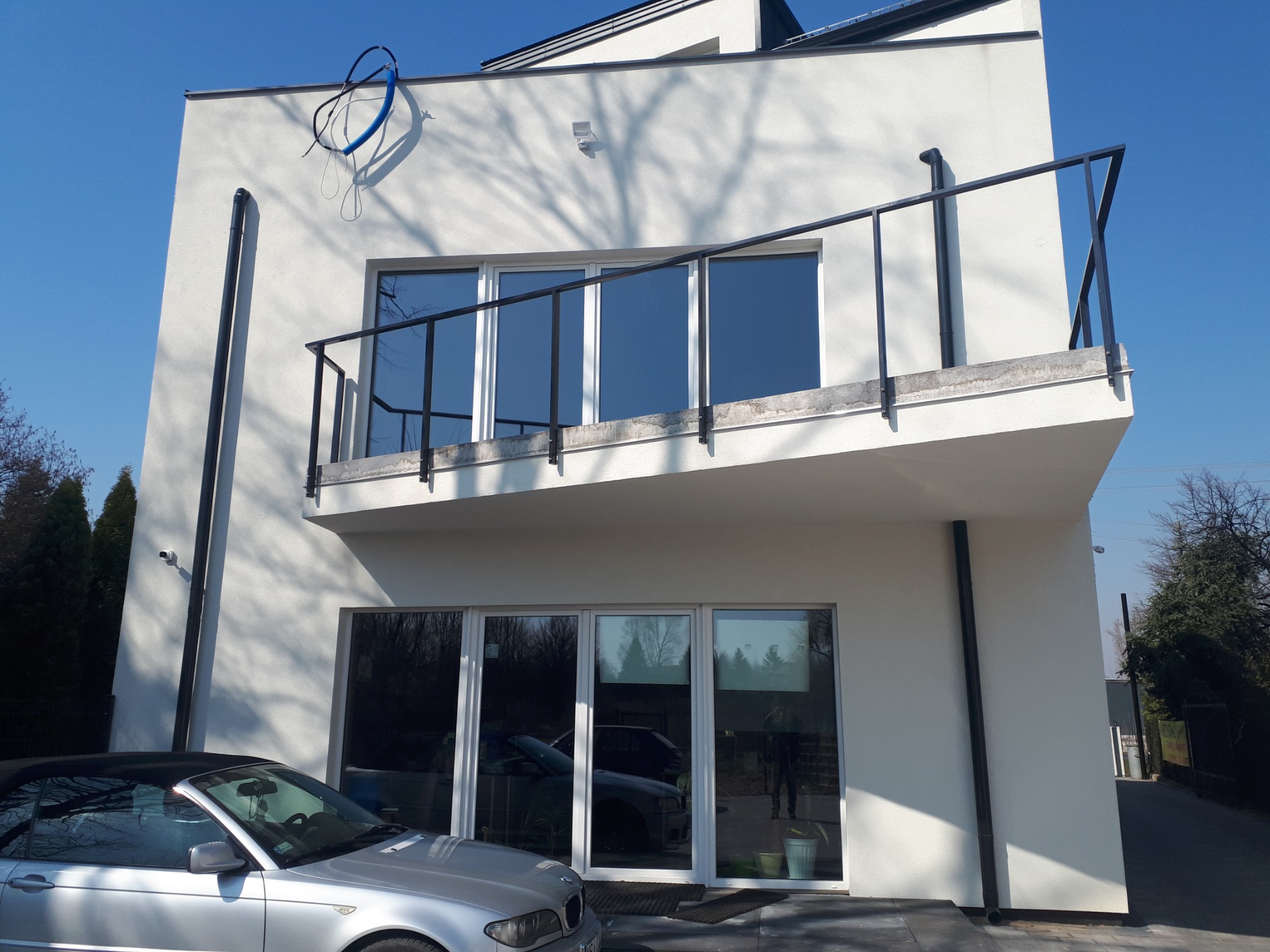 Biała elewacja domu z balkonem i dużymi oknami, cień drzewa na ścianie, srebrny kabriolet zaparkowany przed budynkiem, błękitne niebo w tle.
