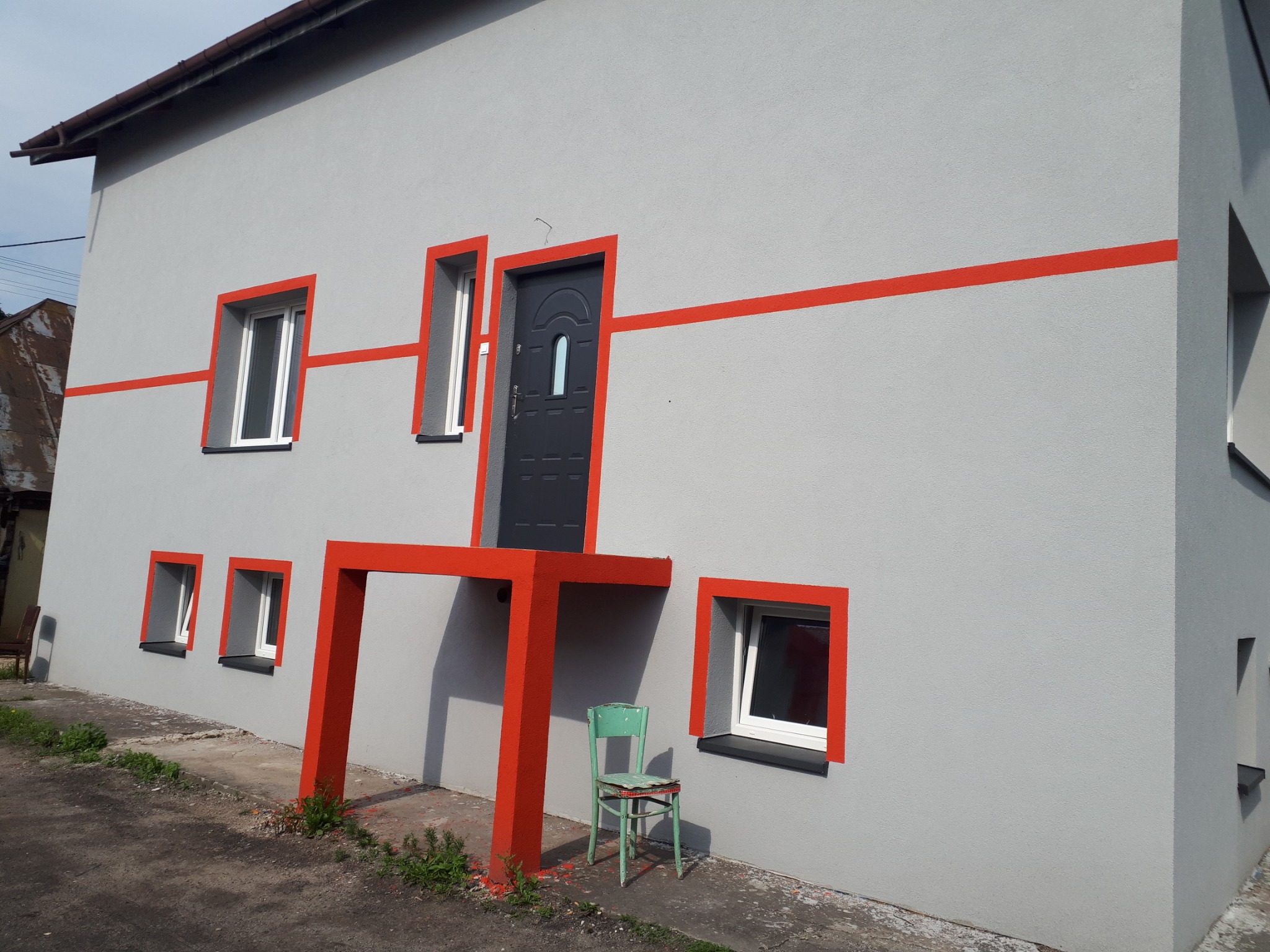 Szara elewacja budynku z pomarańczowymi akcentami wokół okien i drzwi, z widocznym fragmentem chodnika i zielonym krzesłem przy ścianie.