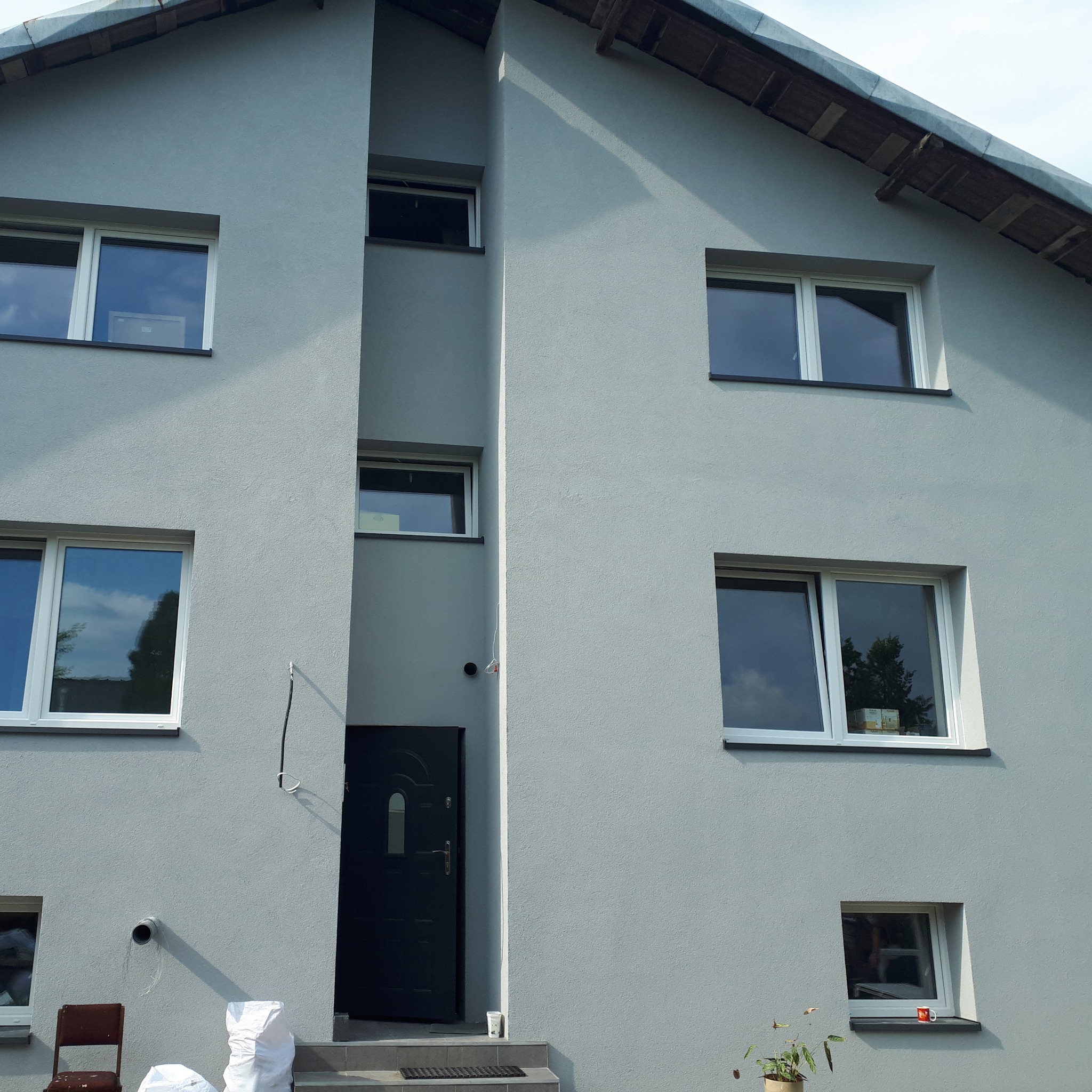 Elewacja domu po tynkowaniu w odcieniach szarości, widoczne białe ramy okienne, czarne drzwi wejściowe, worki z materiałami budowlanymi i krzesło na schodach.