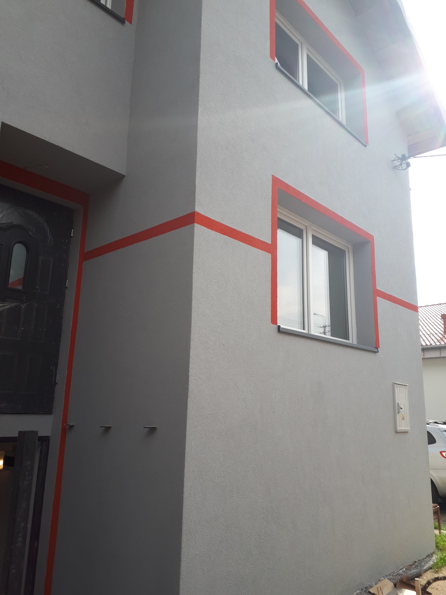 Szara elewacja domu z pomarańczowymi akcentami wokół okien i drzwi, widoczne świeżo pomalowane ściany i nowa stolarka okienna.