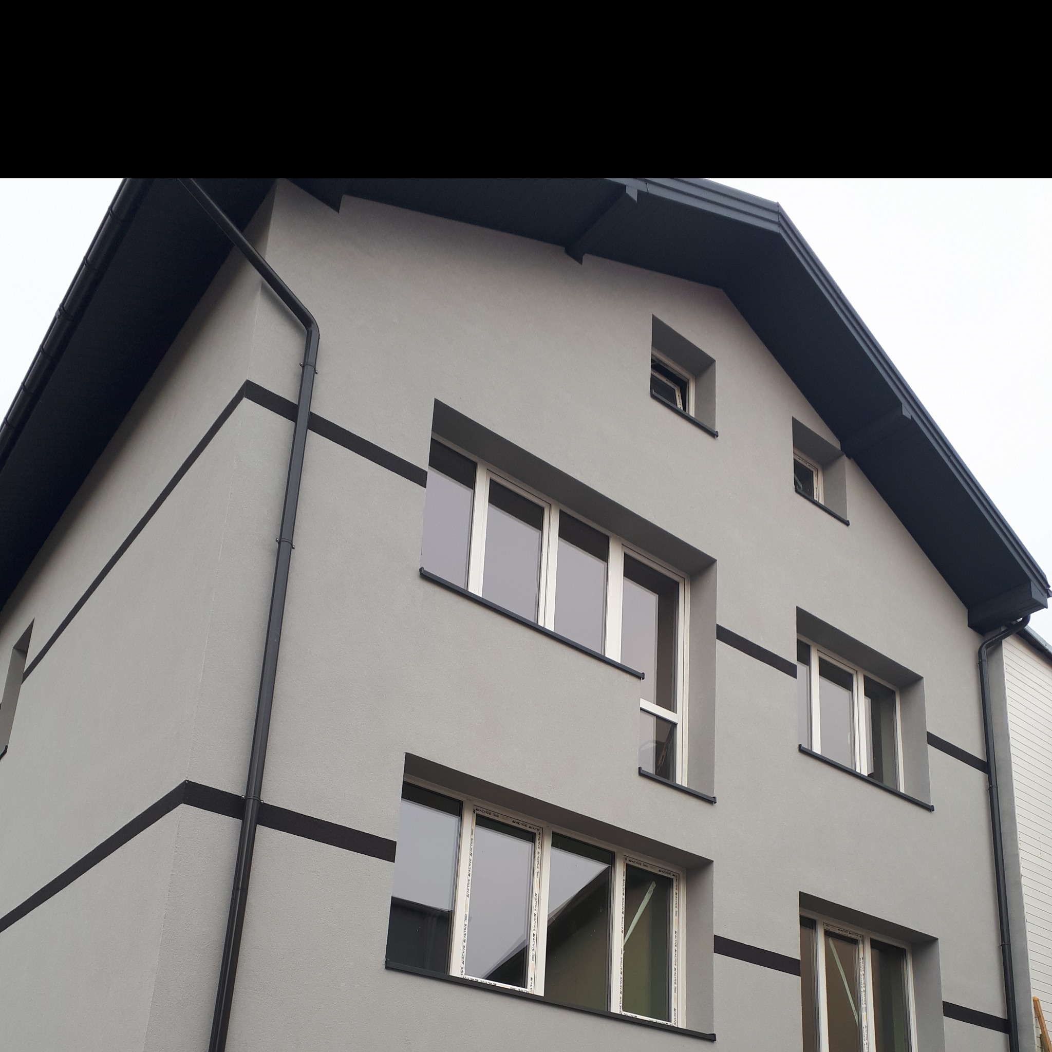 Elewacja budynku mieszkalnego w odcieniach szarości z czarnymi akcentami, widoczne okna PCV z białymi ramami i rynna spustowa.